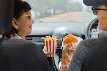 Smiling senior couple enjoying burgers, potato chips , and drinks inside a car during a road trip. Concept of active aging, retirement lifestyle