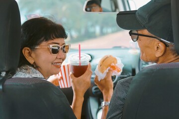 Smiling senior couple enjoying burgers, potato chips , and drinks inside a car during a road trip. Concept of active aging, retirement lifestyle