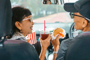 Smiling senior couple enjoying burgers, potato chips , and drinks inside a car during a road trip. Concept of active aging, retirement lifestyle