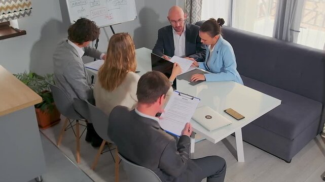 High-angle shot of corporate team in modern office sharing contracts and pointing at data sheets. Professionals in formal attire engaged in active discussion and teamwork with laptops and clipboards.