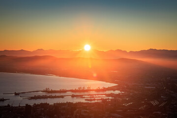 View of the sun ablaze with golden light over misty mountains and the tranquil bay, casting long shadows across the city, Cape Town, Western Cape, South Africa.