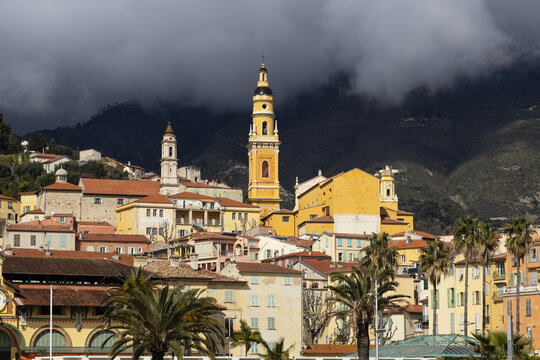 View of ochre and pastel buildings climbing the hillside under a stormy sky, crowned by a striking yellow church tower, Menton, Provence-Alpes-C&Atilde;&acute;te d'Azur, France.