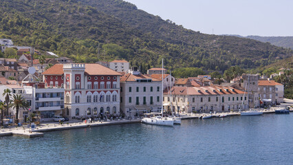 View of aged buildings with red roofs meet the blue waters of the harbor, backed by green hills under a bright sky, Vis, Split-Dalmatia County, Croatia.