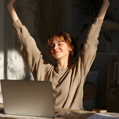 Joyful young woman celebrating success with arms raised in triumph while working on laptop computer at home office desk warm sunlight.
