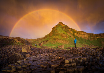 View of a lone figure in a bright blue jacket stands silhouetted against a vibrant rainbow arching over a green hill, Bushmills, Northern Ireland, United Kingdom.