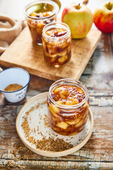 Homemade apple chutney in glass jars with visible chunks of apple, pear, dried fruit, ginger, and spices. Preserved autumn condiment on a rustic wooden background, perfect for seasonal cooking.