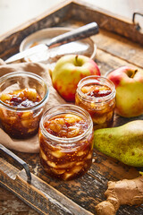 Homemade apple chutney in jars, styled with fresh apples, pear, and ginger in the background. Seasonal preserve with visible chunks of fruit and spices, rustic kitchen setting.