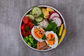 Poke bowl with smoked redfish, eggs, vegetables, edamame beans and rice served on gray table. Healthy balanced meal with white fish. Top view