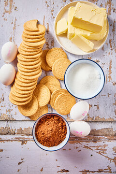 Flat lay of ingredients for traditional Dutch arretjescake, also known as refrigerator cake. Butter, cocoa powder, sugar, eggs, and biscuits arranged on a rustic wooden background.