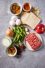 Flat lay of fresh ingredients for traditional South African bobotie on a gray surface. Ground beef, green beans, onion, garlic, eggs, apple, white bread, milk, chutney, raisins, almonds, and spices.
