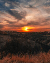 A dramatic sunset with a fiery orange sun illuminating the sky and stratus clouds. In the foreground is a dry, rugged landscape with ravines or embankments, possibly damaged by erosion or fire.