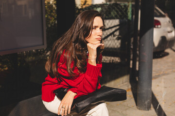 Brunette long hair woman sitting on bus station on bench, waiting for bus, hold chin. Girl wear red sweater, black bag and beige jeans. Sad woman long time waiting bus.