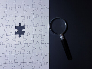 White jigsaw puzzle with a missing piece and magnifying glass on black background. Concept image for problem solving, solution search, research, teamwork, mystery, and analysis.
