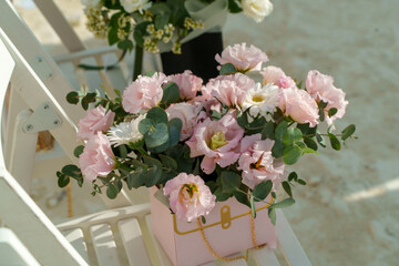 A lovely arrangement of pink flowers in a decorative box sits on a chair near the ocean. Fresh daisies and greenery complement the elegant blooms against the sandy backdrop.