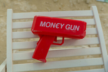 A vibrant red money gun sits on a white chair, surrounded by a sandy area. This playful prop adds excitement to an outdoor gathering, perfect for creating memorable moments.