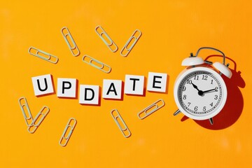 Concept of a timely update or deadline, featuring an alarm clock, paper clips, and letter tiles on a bright orange surface