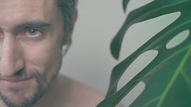A handsome man with expressive eyes stands beside a lush monstera leaf, showcasing a range of emotions from serious contemplation to playful smirk in a soft, calming atmosphere