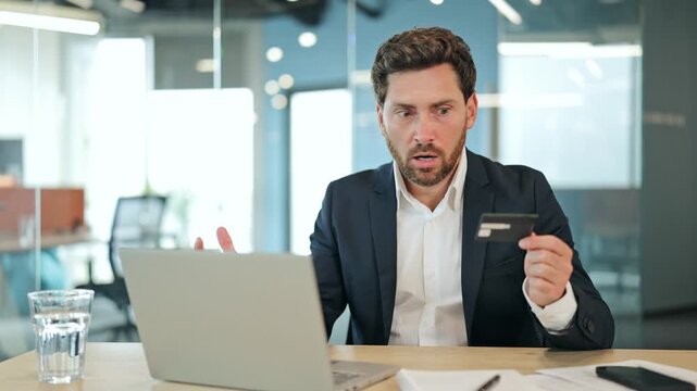 Worried handsome businessman in a professional suit experiences a shocking online payment issue with a credit card on a laptop at an office desk, showing frustration.
