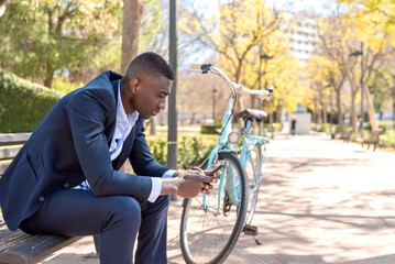 Young African american businessman sitting on bench using mobile phone next to bicycle in city park