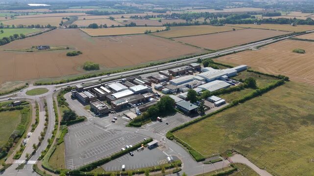Aerial view of industrial buildings with silver roofs surrounded by green and brown fields and a highway cutting through, Nuneaton, England, United Kingdom.