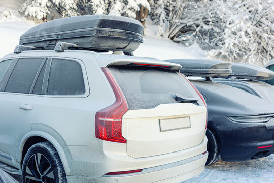 Back view of modern electric car with rooftop box covered frosty snow hoarfrost at alpine street outdoor parking oh cold winter day. Snow-covered SUV roof box parked outdoors in winter conditions