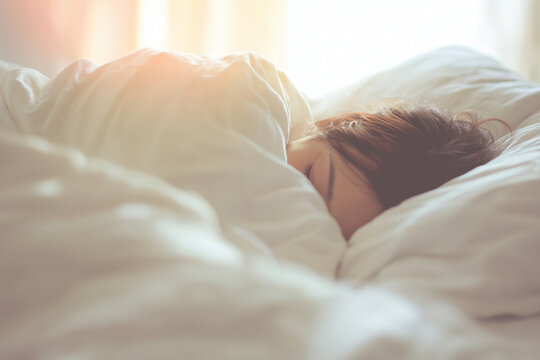 Young person sleeping peacefully under soft white blankets in a sunlit bedroom, cozy morning scene with warm natural light creating a calm and comfortable atmosphere.