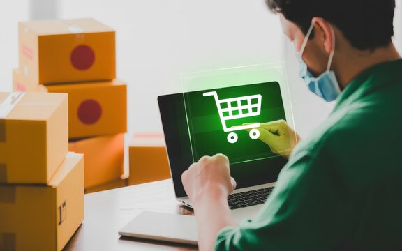 Man wearing a protective face mask orders goods online using a laptop, with a glowing shopping cart icon on the screen and cardboard boxes ready for shipping