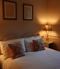 Evening view of traditional English bedroom with lit bedside lamp, carved headboard, floral cushions and shuttered window, concept of home comfort, quiet rest and warm countryside interior