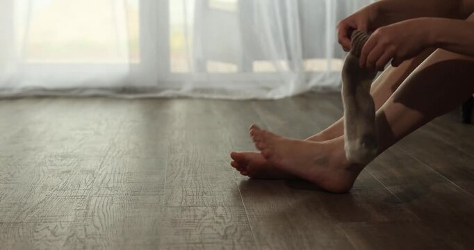 A woman sits on a wood floor with underfloor heating on a bright morning. She rubs her feet and puts on socks to keep warm and comfortable in her home.