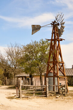 Rustic windmill and wooden fence at Dixie Dude Ranch in the Texas Hill Country, with ranch buildings and trees in the background under a blue sky.