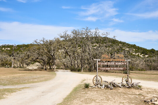 Entrance sign of Dixie Dude Ranch in Bandera, Texas, with a dirt road leading through the Texas Hill Country landscape. Rural western scene with trees, hills, and open sky.