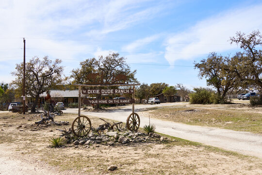 Entrance sign of Dixie Dude Ranch in Bandera, Texas, with a dirt road leading through the Texas Hill Country landscape. Rural western scene with trees, hills, and open sky.