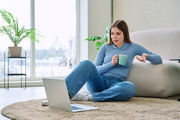 Young relaxed woman with cup using laptop, sitting on floor at home