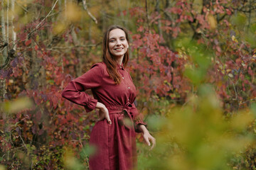 Young woman in a simple red dress smiles naturally among autumn foliage, her genuine expression and...