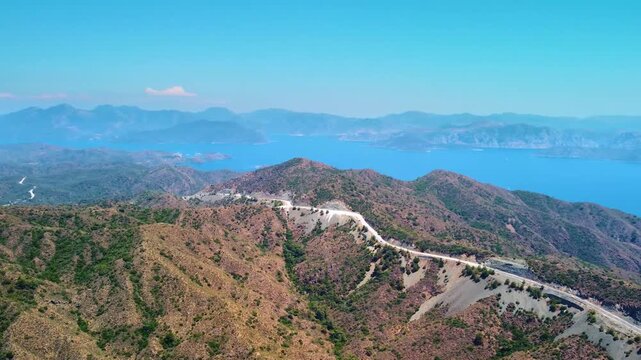 Aerial view capturing the breathtaking journey along a winding coastal mountain road leading to the historic Knidos Antik Kenti in Turkey. The footage showcases stunning panoramic vistas of the Aegean