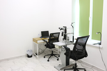 Modern ophthalmologist office interior with slit lamp biomicroscope, desk with laptop, and two chairs, bright and clean medical examination room with white and green design.