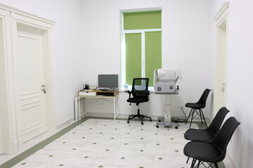 Modern ophthalmologist office interior with slit lamp biomicroscope, desk with laptop, and two chairs, bright and clean medical examination room with white and green design.