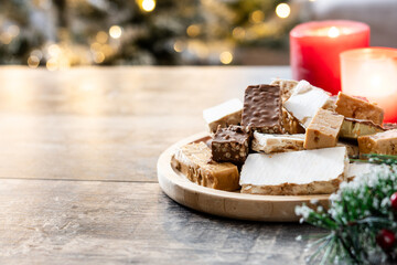 Assorted traditional Christmas nougat on wooden plate and christmas tree background. Copy space