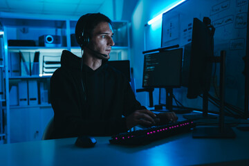 Caucasian male IT support and programmer wearing headset, working night shift on computer screens in dark office.