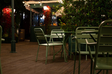 Cozy, dimly lit cafe terrace with metal green chairs and table, surrounded by plants and lights, evokes peaceful, intimate atmosphere perfect for relaxation or quiet conversations under night sky