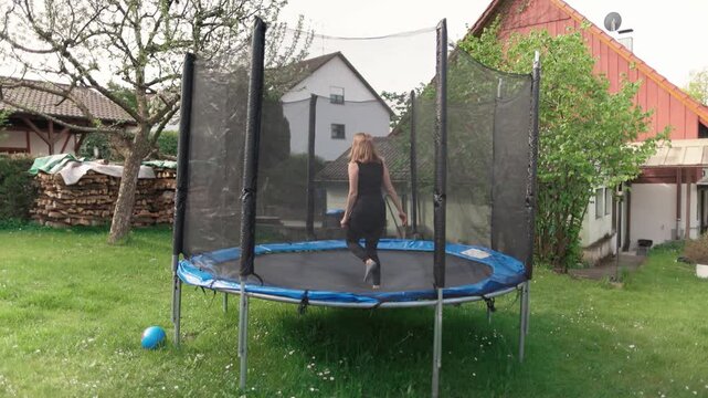 Girl is jumping on a trampoline in her backyard with green grass in the country house