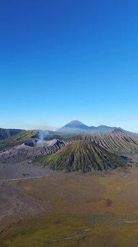 Vertical, aerial: Mount Semeru erupting above Bromo Caldera during the day with blue sky in East Java, Indonesia, establishing drone shot