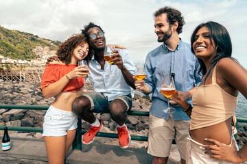 Joyful multicultural friends laughing by the sea on summer vacation in Italy. Diverse group of young people embracing. Friendship and travel concept.