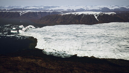 Majestic glaciers extend over rocky terrain, meeting dark waters in a remote Arctic setting. Snowy mountaintops rise in the background under a clear sky, showcasing natures beauty.