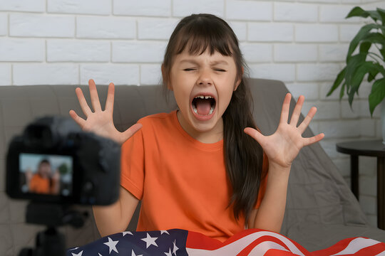 Angry young girl shouting at camera during home video recording, emotional child expressing frustration with hands raised and mouth open, female kid in orange shirt sitting behind American flag fabric