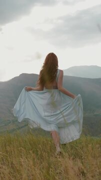 Slow motion vertical video redhead woman running on mountain trail back view dress and hair flowing in wind cinematic lifestyle freedom adventure nature outdoor scenic beauty