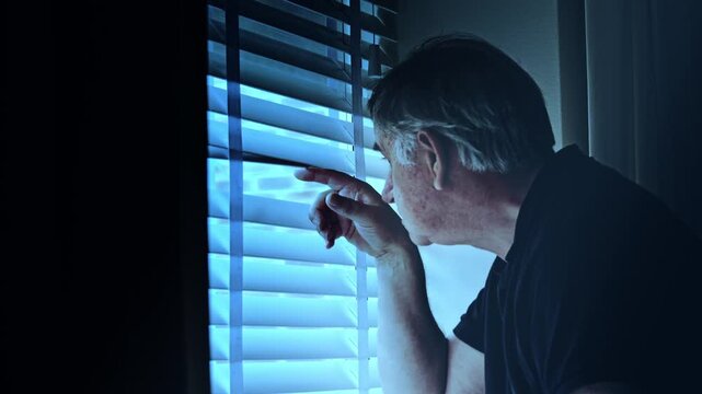 Mature man with grey hair in a dark room looks fearfully out the window through window blinds at night