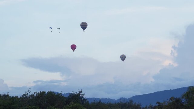 Several large, brightly colored hot air balloons, including patterned solid red designs, gently ascend through wide, partly cloudy sky. Two powered paragliders paramotors also visible upper left,