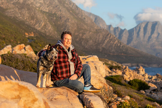 A man sitting outdoors with his Australian Cattle dog (blue heeler), looking at the view, hiking and exploring together, the dog is alert