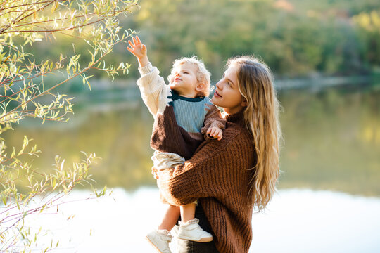 Happy blonde woman holding baby boy wearing knitted sweater over nature lake background close up. Autumn season. Motherhood.